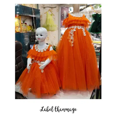 A mother-daughter twinning set in bright orange tulle, displayed on mannequins. Both the gown and the frock feature an off-shoulder neckline with white floral applique details.