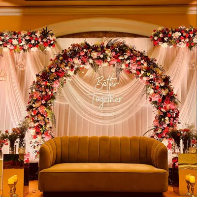 A romantic stage backdrop featuring a lush floral arch of red and pink roses, with soft white drapes and a "Better Together" sign, perfect for couple's photos.