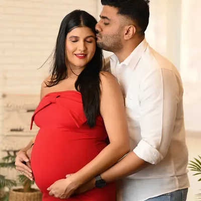A tender moment between an expecting couple, with the partner kissing the mother-to-be's forehead. The woman is wearing a vibrant red dress, adding a pop of color to this studio maternity portrait.