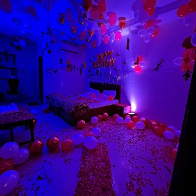 A full room decorated for a birthday surprise, with red and white balloons, a rose petal pathway, and moody blue lighting.