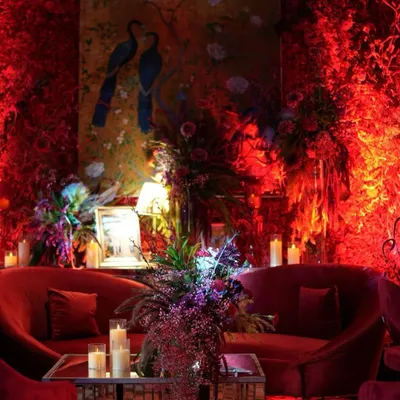 A luxurious lounge area drenched in shades of red. The space features plush red velvet sofas, a mirrored coffee table, and walls covered in red foliage, all illuminated by warm, dramatic lighting. A hand-painted peacock artwork adds a touch of artistry to the passionate design.