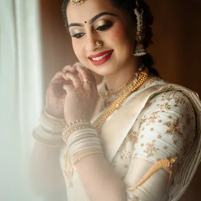 A beautiful bride getting ready in an elegant off-white Kanchipuram saree. Her custom blouse features delicate gold floral embroidery that perfectly matches her traditional gold jewelry.