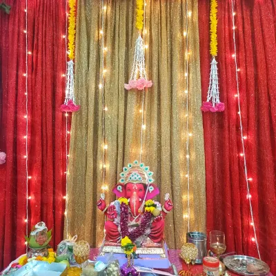 A vibrant home Ganpati mandap featuring a bright red idol. The backdrop combines a shimmery golden curtain with rich red drapes, accented by fairy lights and hanging floral tassels for a classic festive look.