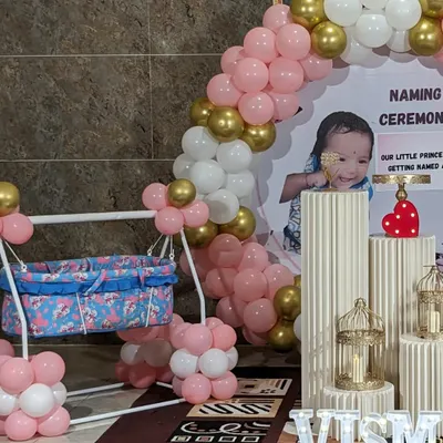 A modern naming ceremony setup featuring a personalized backdrop with the baby's photo. The decor includes a pink, white, and gold balloon arch, a decorated cradle, and illuminated letters spelling the name.