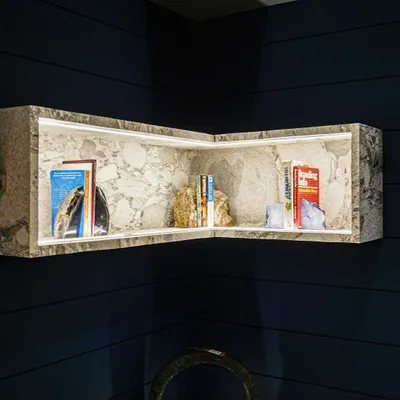 This custom corner bookshelf is crafted from a single piece of stone, with integrated LED lighting that casts a warm glow on the displayed books and objects. Set against a dark blue grooved wall, it stands out as a piece of functional sculpture.