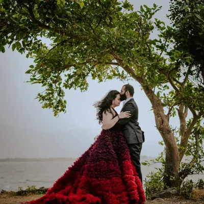 A truly romantic pre-wedding moment captured under a lush green tree by the water. The wind catching her hair and the stunning red ombre gown makes this shot feel like a movie scene.
