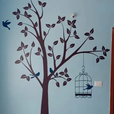 A large tree stencil in dark brown on a light blue wall creates a serene nature scene. The design includes small birds perched on branches and a decorative birdcage, adding a whimsical touch.