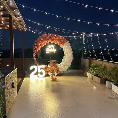 A stunning rooftop terrace party for a 25th birthday, featuring a rose gold and white balloon ring under string lights against the night sky.