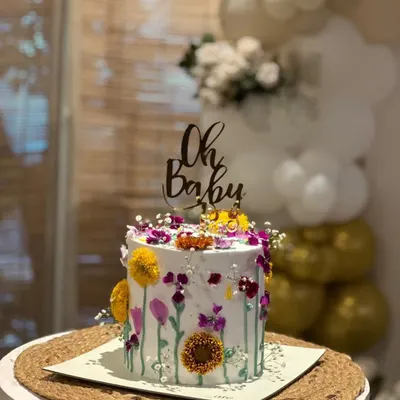 A simple and elegant "Oh Baby" topper in dark wood or acrylic, sitting atop a beautiful wildflower cake. It's the perfect touch for a rustic or boho-themed baby shower.