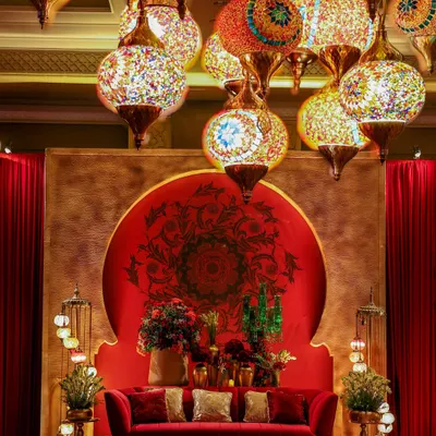 A central seating area designed for a Sufi-themed night. A plush red velvet sofa is set against a circular mandala backdrop, while a cascade of colorful Turkish mosaic lamps provides an intimate and magical glow.
