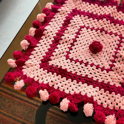This square tablecloth in shades of pink and red is finished with playful pompoms, making it a festive addition to any table setting.