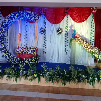 A full stage setup for a naming ceremony with a floral crescent moon swing and a peacock figure. The design uses a mix of blue, white, and yellow flowers with hanging green vines.