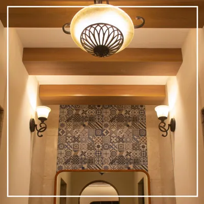 The entrance to the gents' washroom sets a tone of refined elegance. A classic chandelier and traditional sconces illuminate the space, highlighting the intricate patterned tilework that serves as a focal point, framed perfectly by an archway.