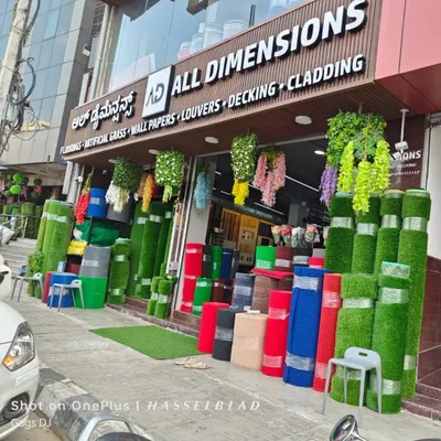 The front of my showroom, "All Dimensions," is stocked with rolls of artificial grass in various colors and textures. You can also see hanging plants and other decor items, giving you an idea of the variety I offer.