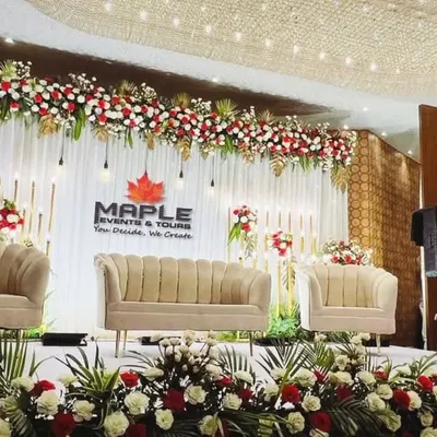 A beautiful stage setup for a formal event, featuring plush seating and a backdrop adorned with red and white floral arrangements. The combination of hanging lights and elegant furniture creates a warm and inviting atmosphere.