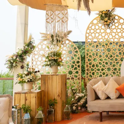 A perfect corner in our rustic-themed decor. This setup features a modern sofa, wooden block tables, and beautiful floral arrangements, all framed by intricate jali-work panels and macrame hangings.