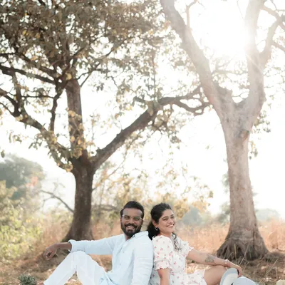 A couple sits back-to-back on a picnic blanket under a beautiful tree, smiling as the sun shines behind them. This shot captures the relaxed and joyful vibe of an outdoor pre-wedding session.