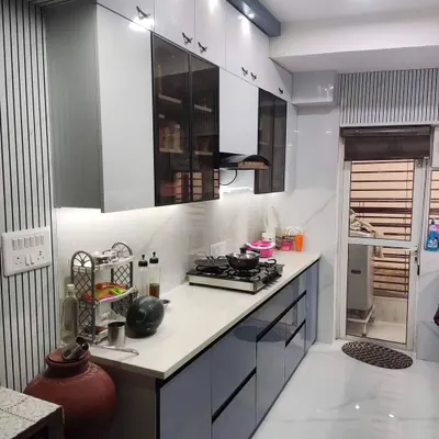 A full view of a compact and modern kitchen we completed in ParasTeria, Noida. The dual-tone design features dusty blue base cabinets and white overhead cabinets with frosted glass inserts. We used sleek, black profile handles and under-cabinet lighting to create a sophisticated and well-lit workspace.
