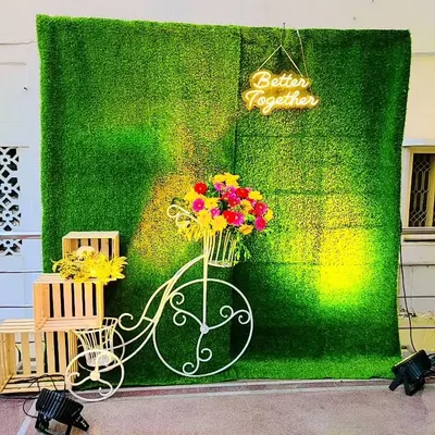 A charming and rustic photo booth perfect for a wedding or engagement party. The setup includes a green turf wall, a "Better Together" neon sign, wooden crates, and a decorative bicycle with flowers.