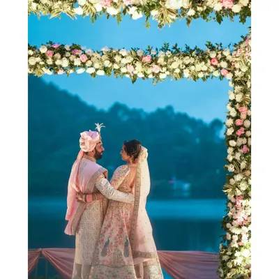 A stunning destination wedding portrait of a couple under a floral arch, with a serene lake and mountains at dusk. This is the magic of a Nainital wedding.