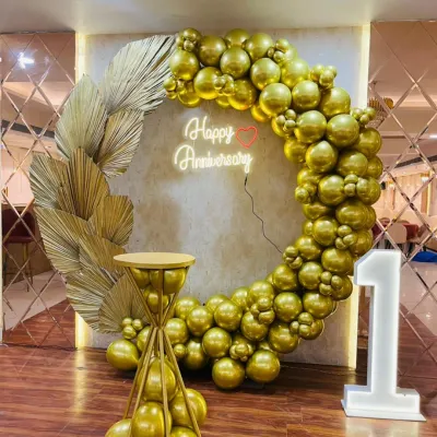 An all-gold theme for a first anniversary, featuring a circular balloon arch made of chrome gold balloons, a neon sign, and a matching cake pedestal. This setup, shown here for a first milestone, can be adapted for any anniversary year.