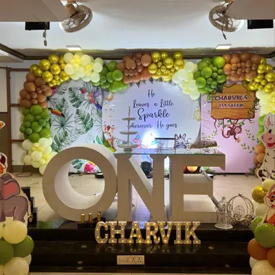 A grand jungle safari themed first birthday stage. The setup includes a large "ONE" prop, a backdrop with tropical leaves, and a balloon arch in earthy tones of green, brown, and yellow.