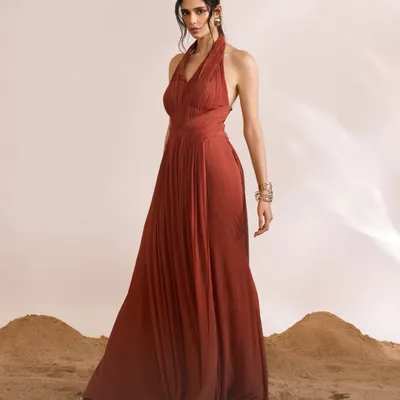 The Luna Dress in a rich rust color, designed with a graceful halter neck and an intricately pleated bodice. The flowing A-line skirt creates a silhouette of timeless elegance.