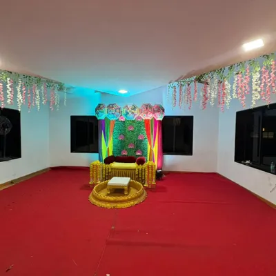 A wide view of a Haldi decoration setup we did in a hall. The space features a colorful stage backdrop, traditional seating, and hanging floral decorations from the ceiling.