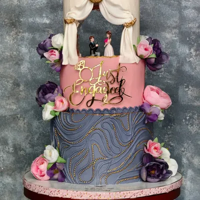 A full shot of the grand three-tiered engagement cake. The design combines a textured grey bottom tier, a smooth pink middle tier with a "Just Engaged" topper, and a top tier featuring a romantic diorama.