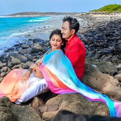A serene pre-wedding portrait of a couple resting on coastal rocks. The vibrant, multi-colored dupatta flows in the breeze against the backdrop of the blue sea, creating a picturesque and romantic scene.