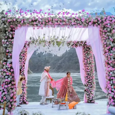 A beautiful moment from a riverside wedding in Rishikesh, with the couple under a mandap lavishly decorated with pink and white flowers. The natural landscape of the river and hills provides a truly unique setting.
