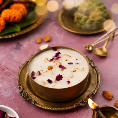 A festive shot of kheer for Navratri. The warm lighting from the diyas and the rich brass bowl create a traditional, celebratory mood, highlighting the creamy texture and delicate rose petal garnish.