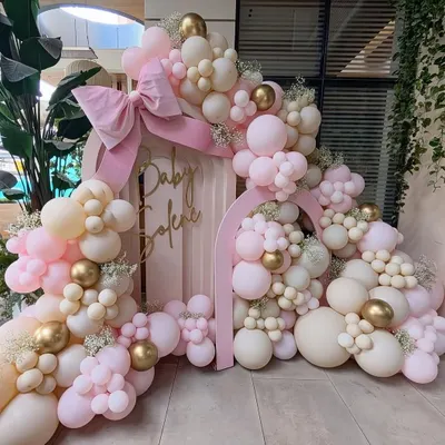 An elaborate and beautiful baby shower setup. The double arch backdrop is covered in an organic arrangement of pink, cream, and gold balloons, with a large pink bow as the finishing touch.