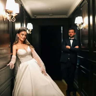 A newlywed couple poses in a classic, wood-paneled hallway. The bride, in her stunning white gown, leans against the wall while her groom stands proudly in the background.