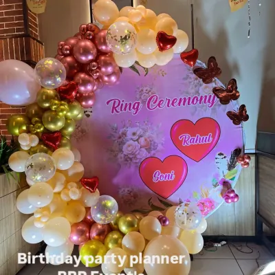 A personalized ring ceremony backdrop with a custom printed floral design featuring the couple's names, 'Rahul' and 'Goni'. The ring is adorned with a balloon arch in shades of pink, peach, and gold.