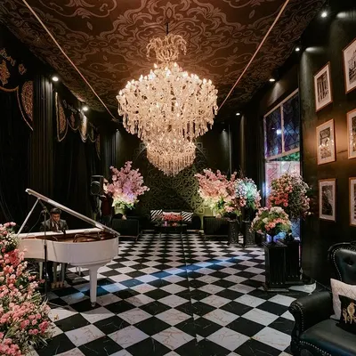 The entrance to a chic pre-wedding event, featuring a grand white piano on a black-and-white checkered floor. The room is filled with crystal chandeliers and soft pink floral arrangements, creating a stunning contrast with the dark, patterned ceiling.