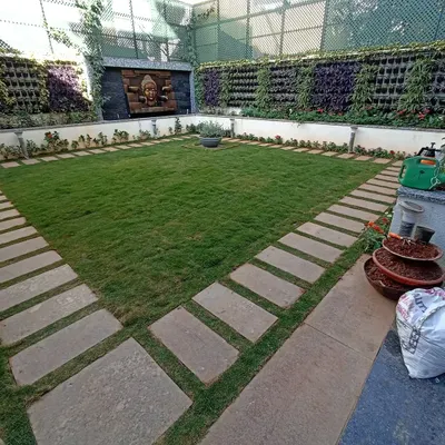 A formal courtyard garden featuring a central lawn bordered by stone pavers. The space is enclosed by lush vertical gardens and a serene Buddha water feature, creating a private and symmetrical oasis.