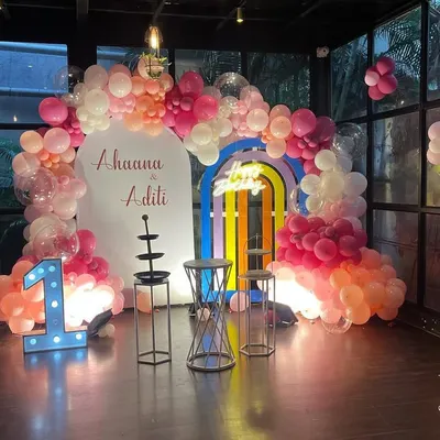 A beautiful pastel-themed first birthday party, featuring a large balloon arch, a rainbow-colored door prop, and a prominent light-up number '1'.