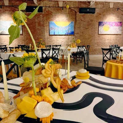An art gallery event where the tablescape itself is a work of art. This centerpiece is crafted from an assortment of cheeses, breads, and sunflowers, set against an exposed brick wall and modern paintings.