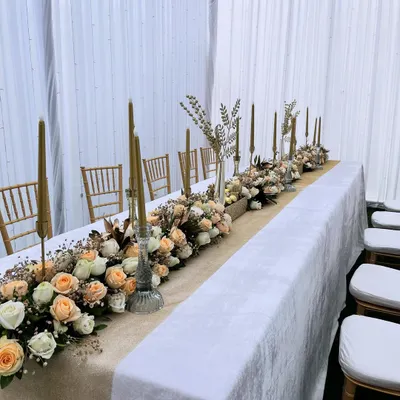 A full view of a long dining table dressed in an elegant floral arrangement. The design features a burlap runner topped with a dense mix of peach and white roses, accented with tall golden candles for a romantic and sophisticated dinner setting.