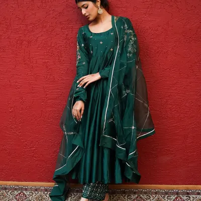 A moment of quiet grace. The deep emerald of the anarkali against the bold red backdrop creates a visually rich and sophisticated portrait.
