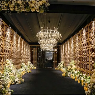 We call this a pre-party runway. A dramatic entrance tunnel with golden geometric walls, a grand chandelier, and a carpet of white florals leading guests to the Sangeet.