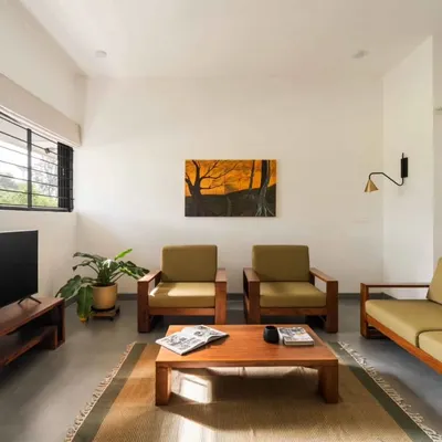 The living area is arranged to feel open and connected, with simple teak wood furniture and large windows that bring in ample natural light. The space is designed for family interaction and relaxation.