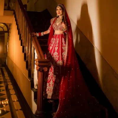 A stunning portrait of the bride on a grand wooden staircase. The dramatic lighting highlights the intricate details of her red lehenga and the elegance of her pose.