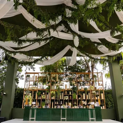 The statement bar from our 'Green Haven' 50th anniversary party. The deep green bar and gold shelving filled with foliage and candles created a sophisticated focal point, set against a ceiling of draped white fabric and hanging ferns.
