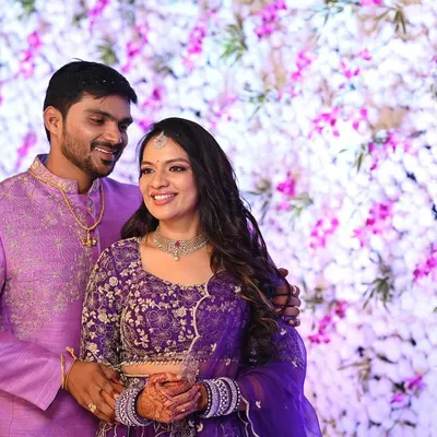 A beautiful couple shot from a wedding reception. The bride's makeup is soft and glamorous, with loose waves and a diamond necklace that perfectly complements her purple lehenga.