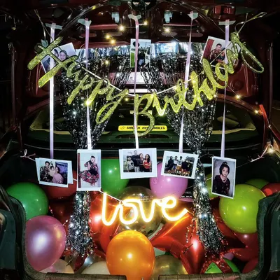 A fully decorated car trunk for a birthday surprise, complete with balloons, a photo garland, fairy lights, and a 'love' neon sign.
