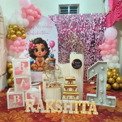 This first birthday setup for Rakshitha features a pink shimmer wall and a Moana-inspired baby character backdrop. The decor is completed with a marquee '1', 'BABY' blocks, and light-up letters for the name.