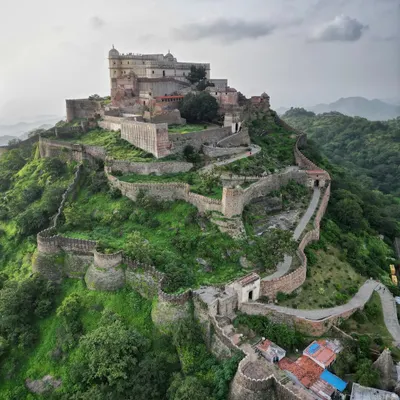 A majestic view of Kumbhalgarh Fort in Rajasthan, a UNESCO World Heritage site. The drone captures the main palace complex perched atop a hill, with its formidable fortification walls snaking through the green landscape.