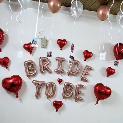 A classic 'BRIDE TO BE' room decoration featuring rose gold foil letters, red heart balloons, and photos of the bride with her friends hanging from the ceiling.